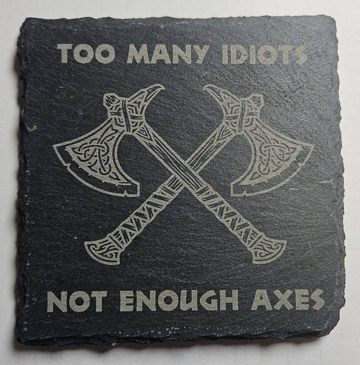 Slate coaster with crossed axes and text 'Too Many Idiots Not Enough Axes' on a grey background